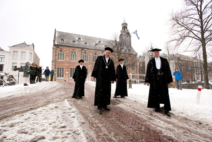 A group portrait of Carel Stolker, Hester Bijl, Annetje Ottow and the beadle, all in academic dress, in front of a snowy Academy Building.