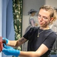 Een jonge vrouw in een zwart shirt en blauwe handschoenen riicht haar blik op een object dat ze aan het  bewerken is. Op de achtergrond een muurschilderij van twee vrouwen in 19e-eeuwse kleding.