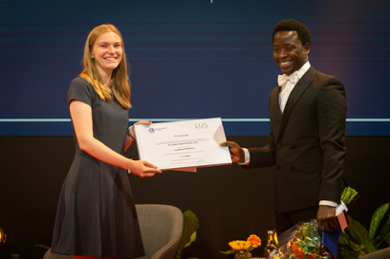 A smiling Ayo Adedokun holds a large bunch of flowers and receives the prize from the chair of the Leiden University Student Platform.