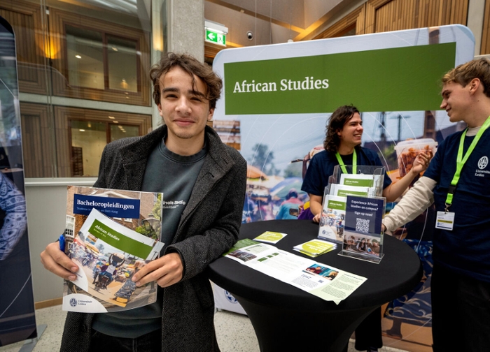 Een jongen met een lange grijze jas en bruin haar, staat voor een banner van African Studies.