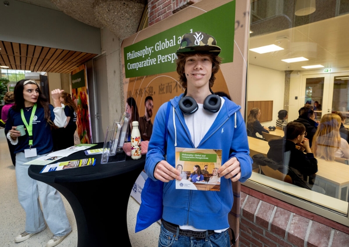 Een jongen met pet op en koptelefoon om zijn nek laat een folder van de opleiding Philosophy zien.