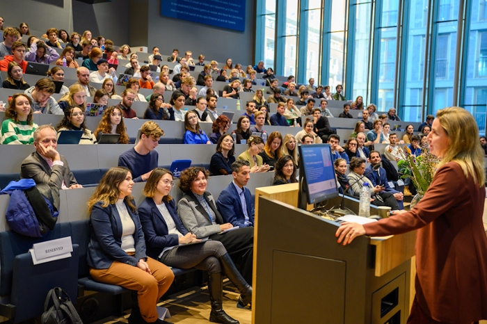 You are looking out on a packed lecture hall, with young students and middle-aged people. The lecturer at the front right of the photo is Frederica Mogherini. She has blond hair and is wearing a brown jacket.