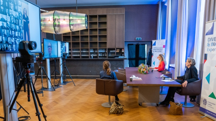 Drie mensen zitten aan een tafel voor een groot scherm met daarop deelnemers aan het symposium