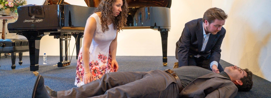 Male student lies on stage, male and female student bend over anxiously with piano in background.