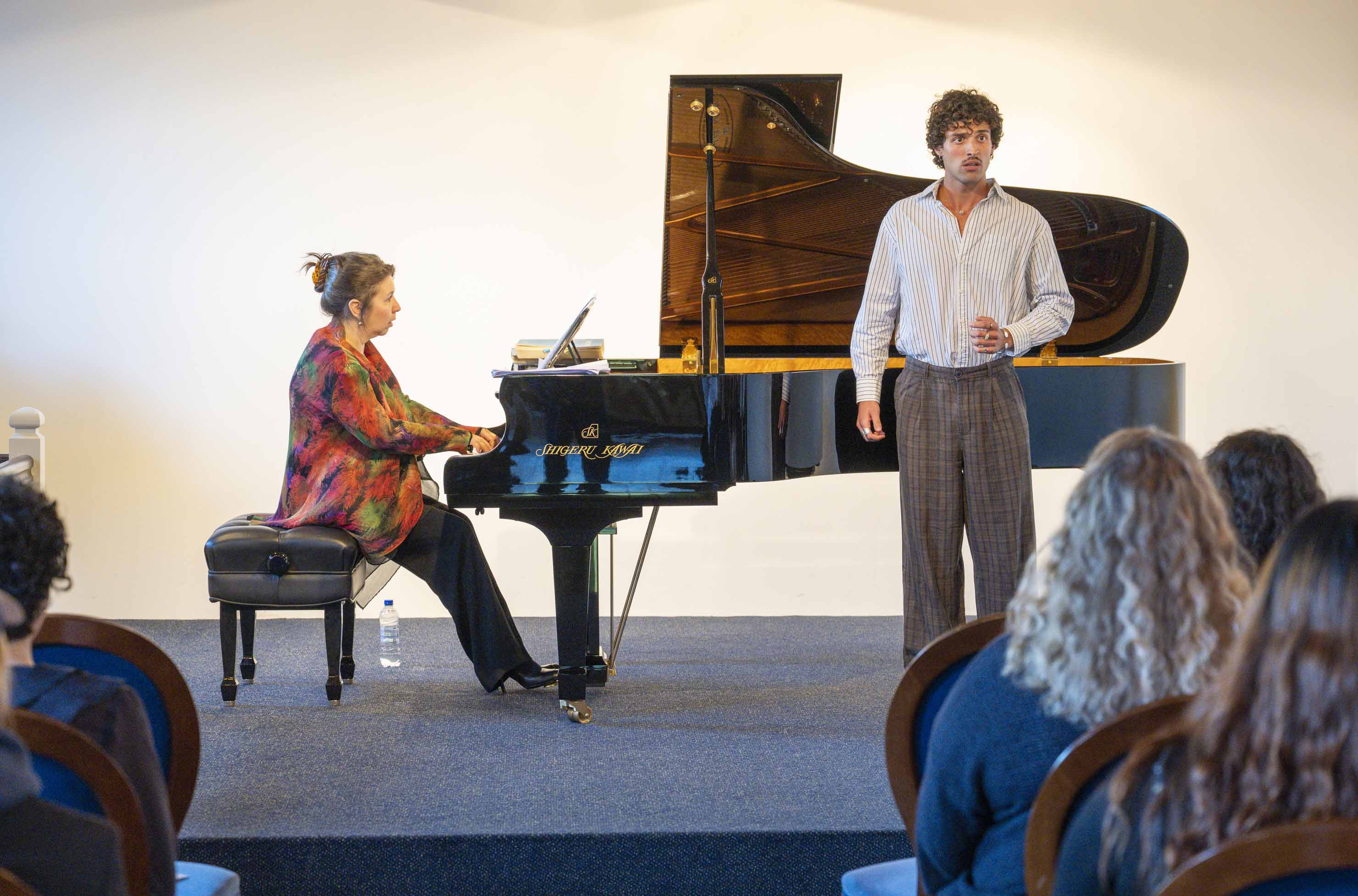 Singer looks into the hall with intense gaze. Behind him, a woman in a red dress plays a grand piano.
