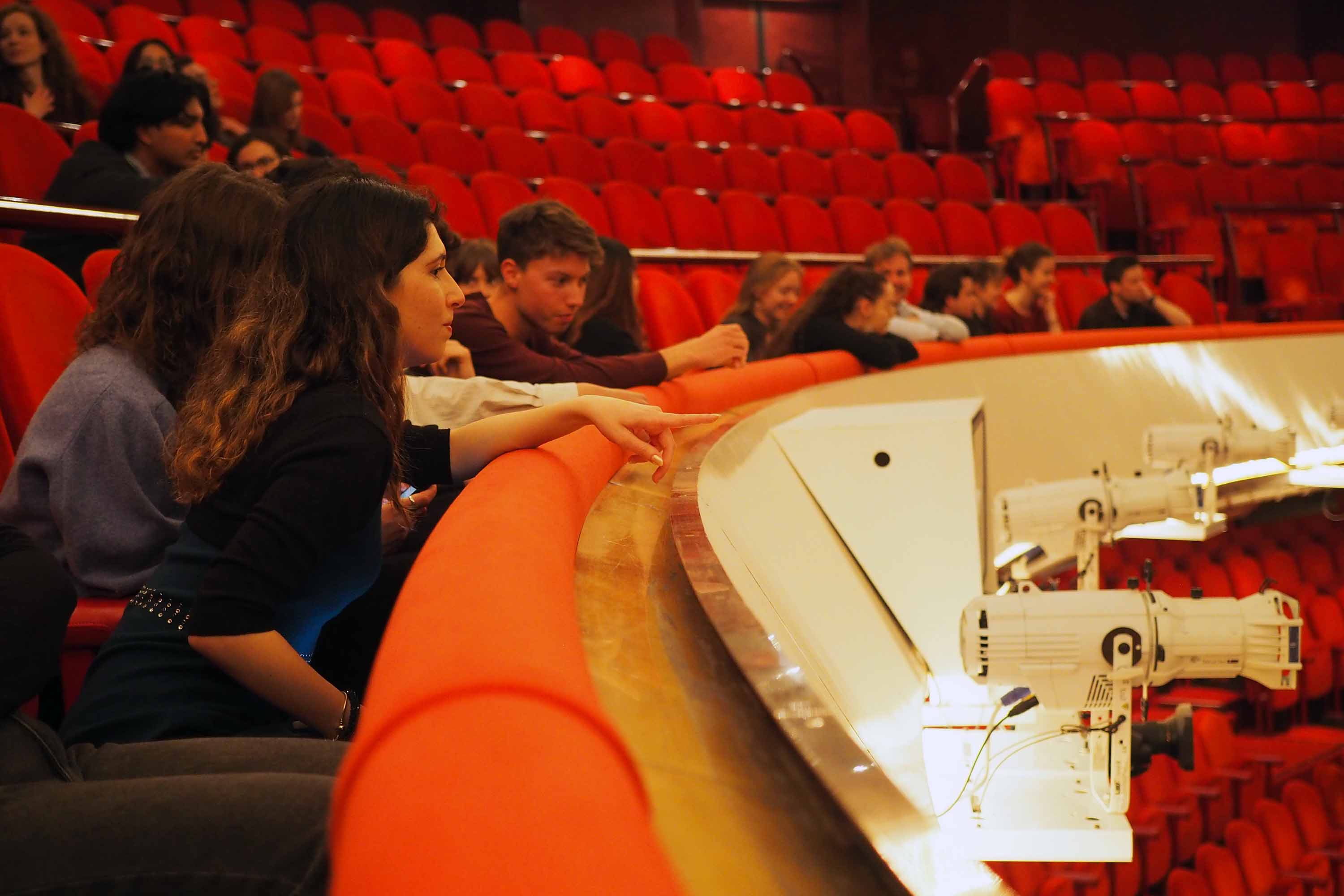 In an opera hall full of red chairs, a student points towards the stage.