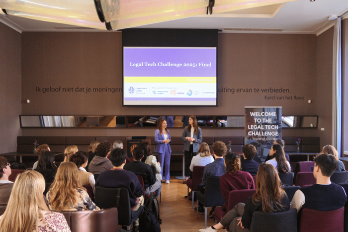 Een groep mensen zitten in een zaal, en kijken naar twee sprekers voor een scherm.